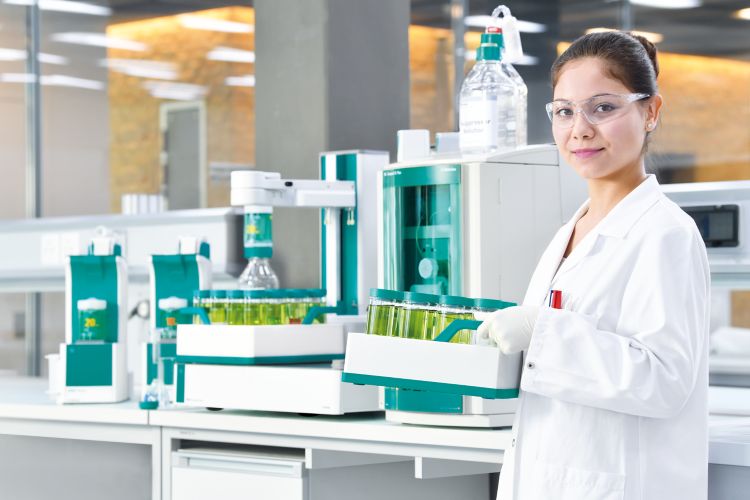 Mulher no laboratório segurando um suporte com diversas amostras verde fosforescente. Ao fundo, na bancada, destaca-se um equipamento e outras amostras.