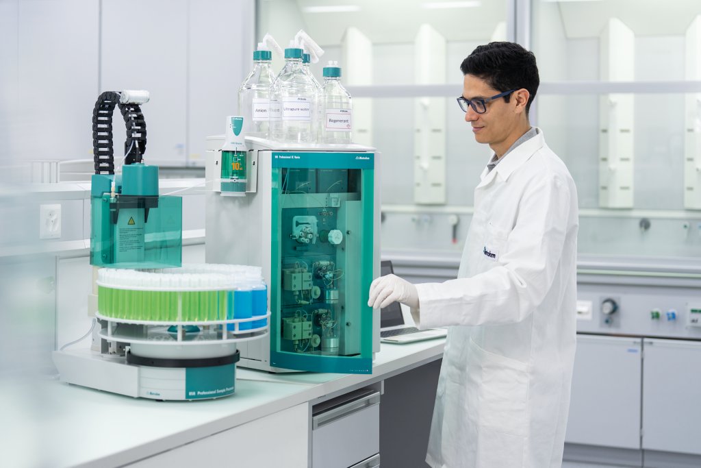 Männlicher Labortechniker steht neben einem offenen Ionenchromatographen und einem vollen Autosampler.