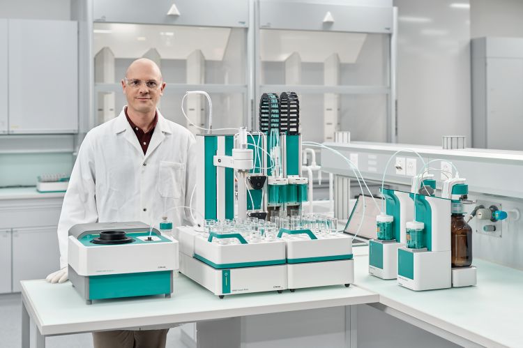 Integrate all your QC analytics on one platform: Lab manager standing behind a OMNIS NIR Analyzer, OMNIS Sample Robot, and OMNIS Titrator. The OMNIS Software is running on a laptop.
